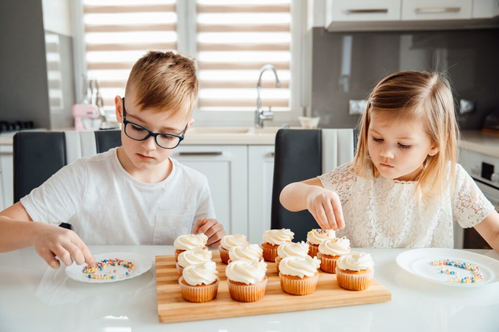 Two children decorate cupcakes by sprinkling icing balls on them